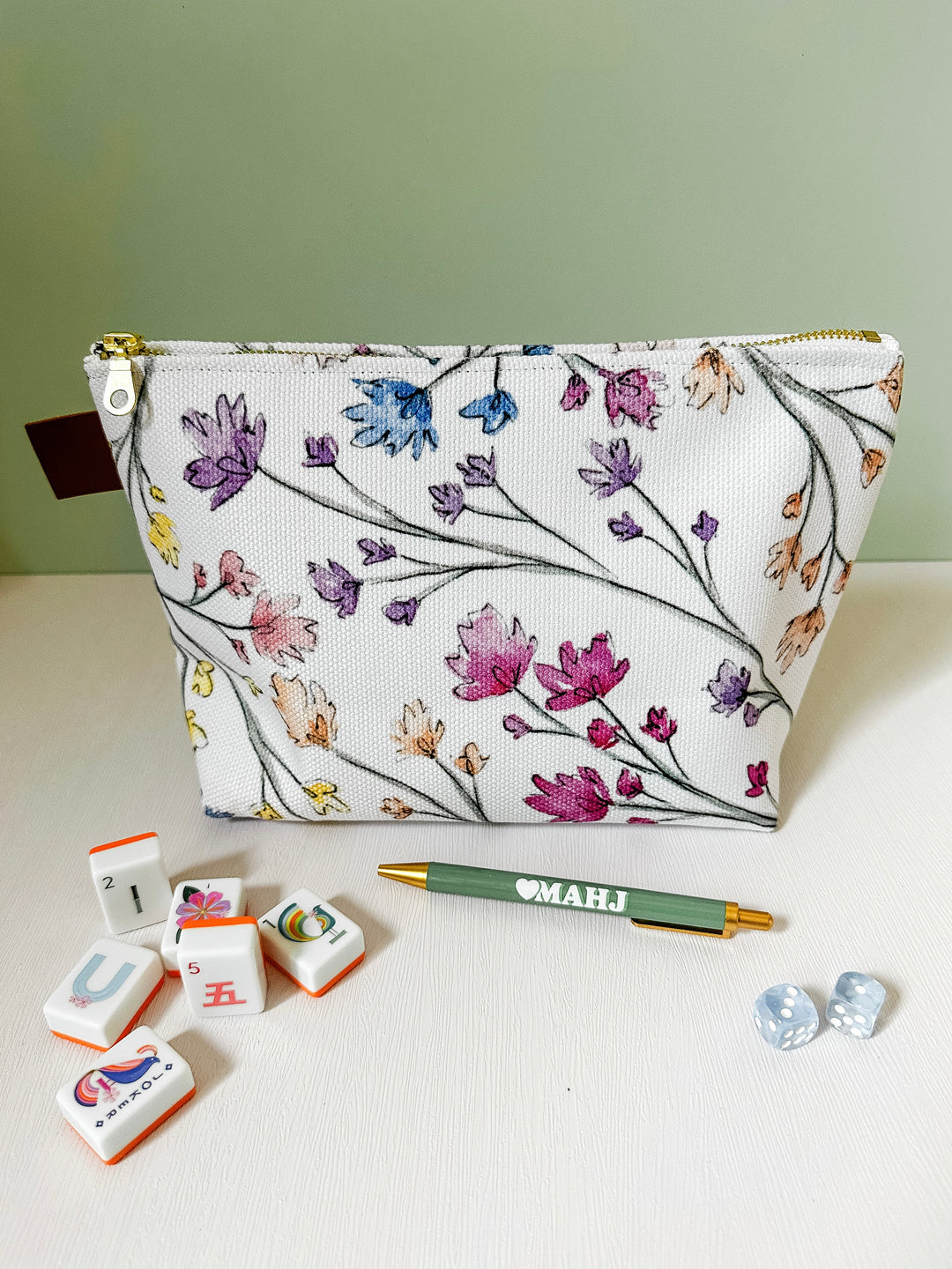 A white zippered pouch with a colorful floral vine pattern, displayed on a white surface with a green pen, mahjong tiles, and two small clear beads in front of it. The background is a soft sage green wall.