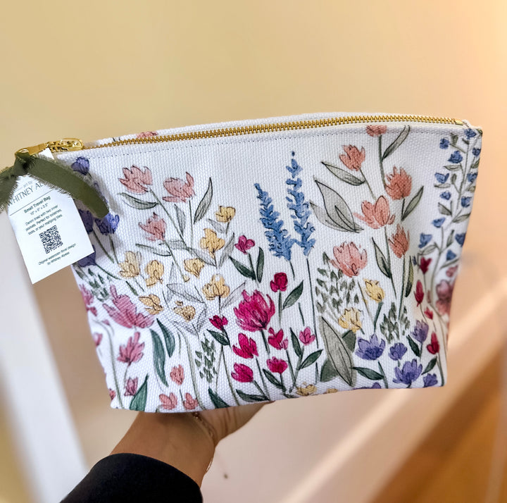 A white zippered pouch with a colorful embroidered floral pattern, held by a hand against a beige background.