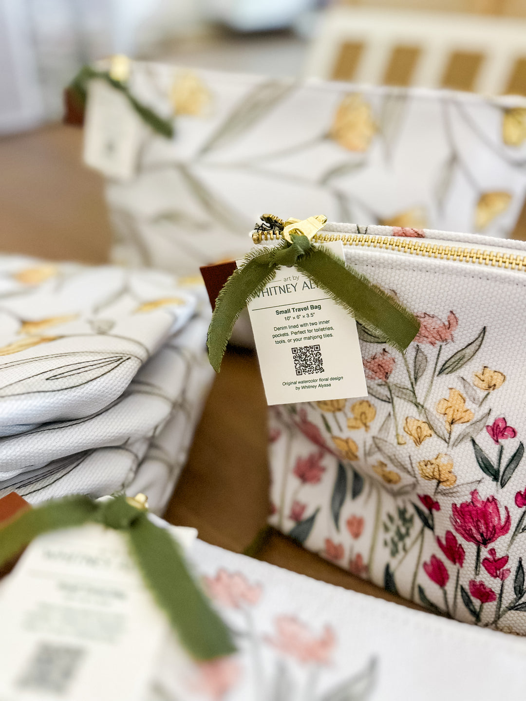A close-up of a white zip pouch with a colorful floral print, tied with an olive green ribbon and a paper tag that reads Whitney Apparel. Other matching pouches are stacked in the background on a wooden surface.